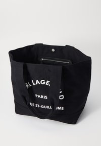 Black canvas tote bag with sturdy straps, featuring large white text on one side and a zippered compartment visible inside.