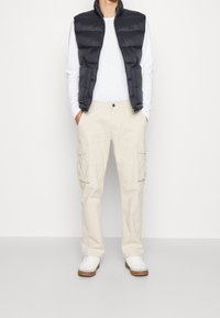 Svart pufferväst, vit långärmad skjorta och beige cargobyxor. Vita sneakers med bruna sulor. Enkelt дизайн med praktiska fickor.