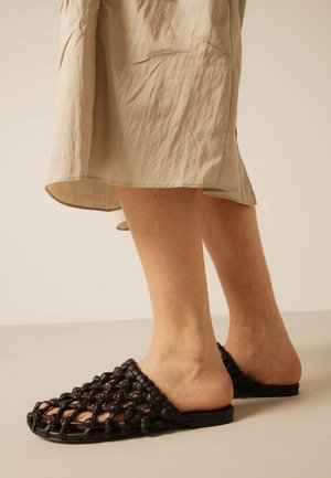 Jambes portant une jupe beige et des sandales mules tissées noires à bout ouvert avec semelles plates sur un fond de sol neutre.