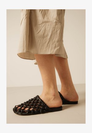 Beine mit beigem Rock und schwarzen offenen gewebten Mule-Sandalen mit flachen Sohlen auf neutralem Bodenhintergrund.