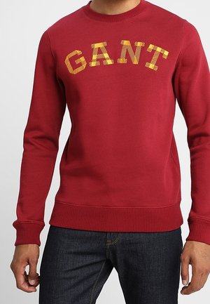 Mężczyzna ubrany w czerwony sweter z okrągłym dekoltem z żółtym wzorzystym tekstem "GANT" na klatce piersiowej i ciemnoniebieskie dżinsy.