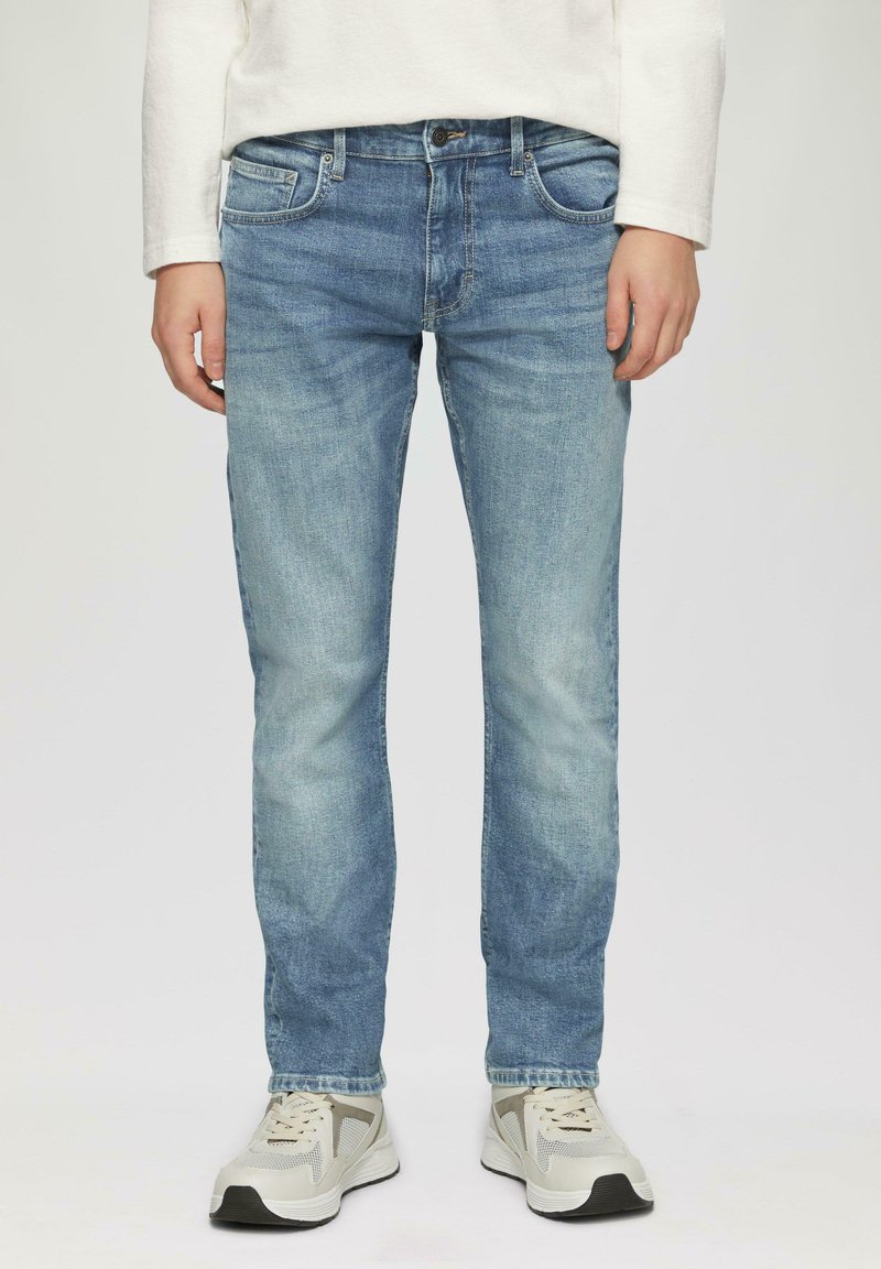 Jeans en denim bleu clair avec une coupe slim, un design à cinq poches et une décoloration subtile. Portés avec des chaussures de sport de teinte neutre.