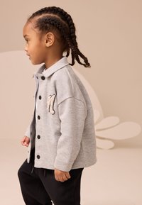 Chaqueta de forro polar gris con botones negros y cuello, con una "M" bordada en beige en el pecho izquierdo, combinada con pantalones negros.