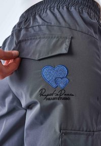 Pantaloni cargo grigi con vita elastica. Tasca posteriore decorata con cuori ricamati blu e il testo "Rayots de Paris HEART STUDIO."