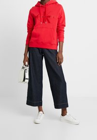 Felpa rossa con cappuccio e logo, abbinata a pantaloni denim larghi scuri e sneakers bianche. Tieni in mano una piccola borsa argento.