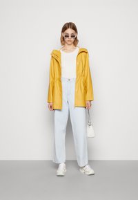 Jachetă galbenă impermeabilă cu talie regulabilă, purtată împreună cu un top texturat alb și blugi evazați de culoare albastru deschis. Accesoriile includ ochelari de soare mici și o geantă albă.