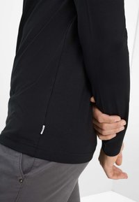 Schwarzes Langarmshirt aus weichem Material mit abgerundetem Saum und einem Etikett an der Seite, kombiniert mit grauen Hosen mit einer Vordertasche.