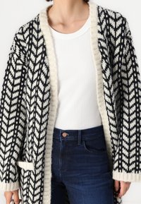 Woman wearing a black and white patterned knit cardigan over a white ribbed top with dark blue jeans, standing against a white background.
