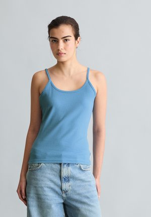 Jeune femme aux cheveux foncés attachés porte un débardeur bleu côtelé et un jean bleu clair, debout devant un fond gris uni.