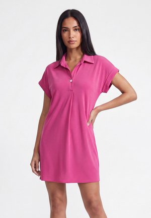 Kvinde med langt mørkt hår iført en pink polo-kjole med korte ærmer, stående med den ene hånd på hoften mod en ensfarvet hvid baggrund.