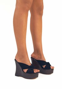Gambe che indossano sandali con zeppa annodata in tessuto nero e plateau scuro su uno sfondo bianco.