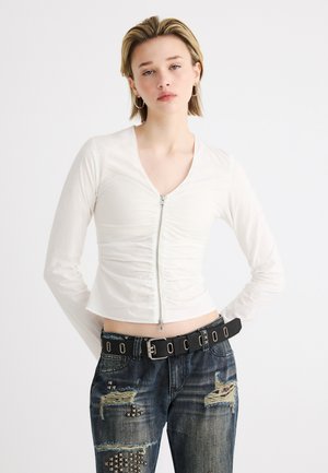 Ung kvinde med kort blondt hår iført en hvid ruchet trøje med lynlås, slidte jeans med nitter og et sort bælte med sølvøjer.