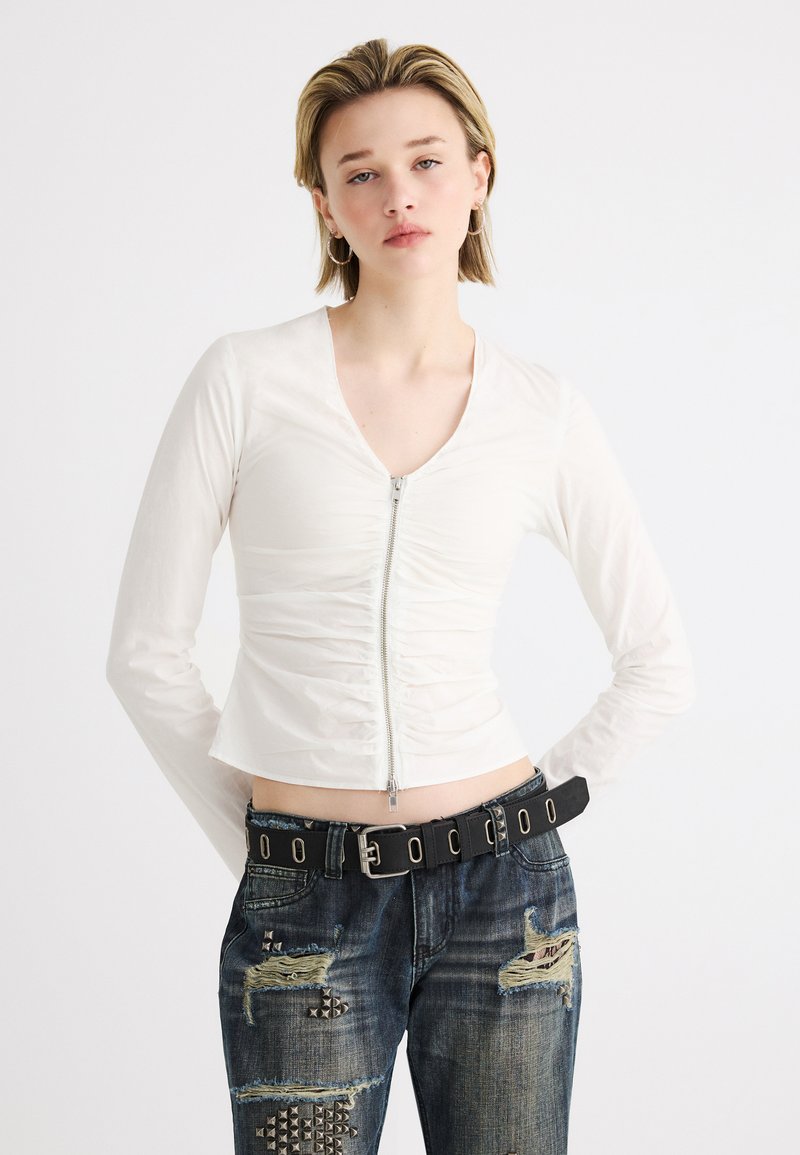 Jeune femme aux cheveux blonds courts portant un haut blanc froncé à fermeture éclair, un jean déchiré clouté et une ceinture noire avec des œillets argentés.