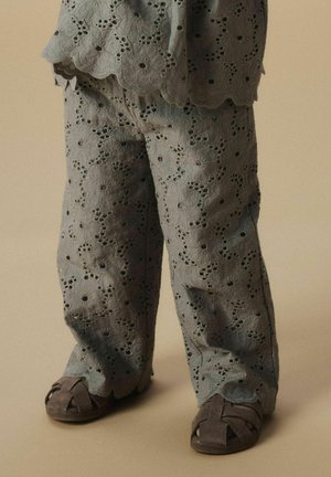 Grijze oogjesstofoutfit met bloemmotieven, gegolfde randen en wijd-uitlopende broeken, gecombineerd met donkergrijze sandalen met kruisbanden.