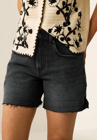 Svart denimshorts med fransade fållar, med en klassisk femficksdesign och bältesloopar. Tyget har en slät, något texturerad finish.