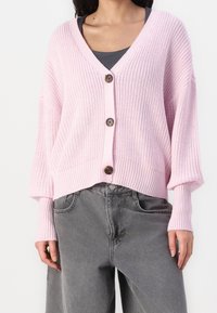 Světle růžový pletený svetr na knoflíky s výstřihem do V, volnými rukávy, třemi hnědými knoflíky na přední straně a zkráceným lemem, doplněný šedými kalhotami.