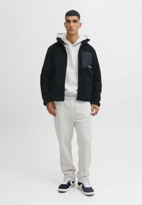 Fleecejakke i sort med en forlomme; grå hættejakke under; lysegrå sweatpants; marineblå sneakers med hvide detaljer.
