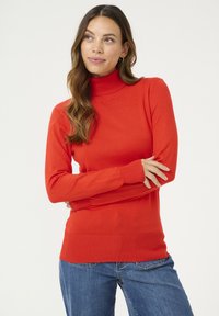 Röd polo-tröja med långa ärmar, ribbade detaljer vid ärmslut och fåll, i kombination med blå jeans, som visar en jämn stickad textur.