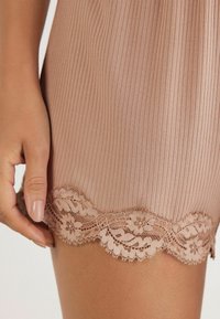 Roze ribbelstof met een scalloped kantzoom. Zachte textuur aangevuld met een delicate bloemmotief van kant. Hand die dichtbij de zoom rust.