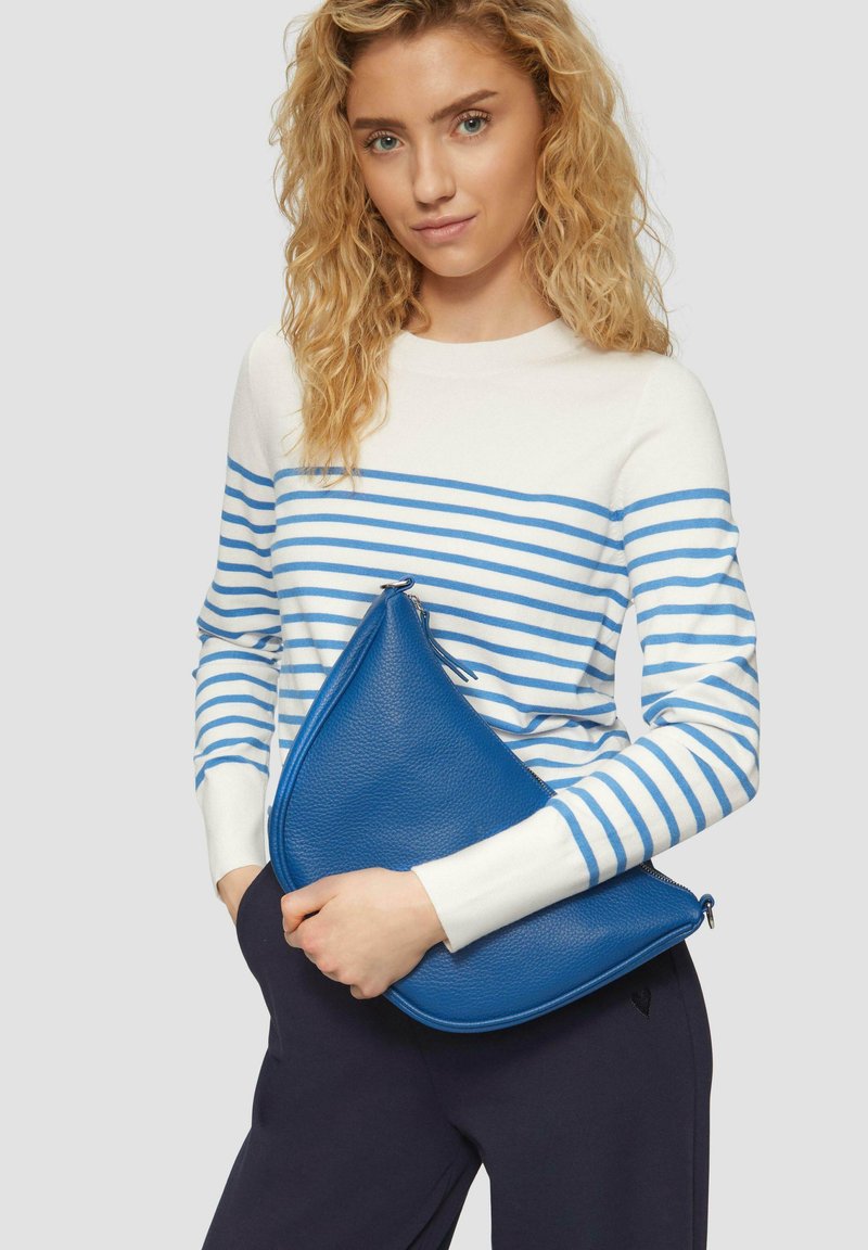 Blaues, dreieckig geformtes Clutch-Bag aus Leder mit Reißverschluss, gehalten gegen einen weiß-blau gestreiften Pullover, der auf einer marineblauen Hose liegt.