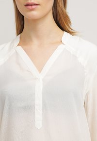 Žena se světlou pletí a blond vlasy má na sobě bílou texturovanou blůzu s výstřihem do V a zapínáním na knoflíky.