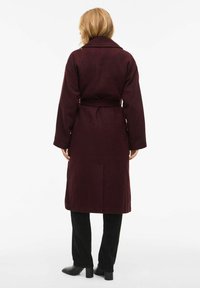 Burgundy coat with a belted waist, long sleeves, and a collar, made from textured fabric. Worn over black pants and heeled boots.