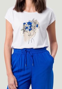 Camiseta blanca con un dibujo lineal de un rostro adornado con acentos florales azules. Combinada con pantalones anchos azules, que muestran un estilo y ajuste relajados.