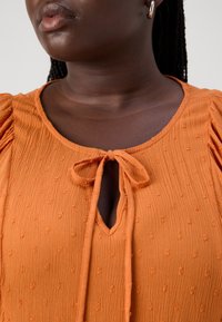 Personne à la peau foncée portant un chemisier orange texturé avec un nœud noué à l'encolure et une petite ouverture en forme de trou de serrure, ainsi que des boucles d'oreilles argentées.
