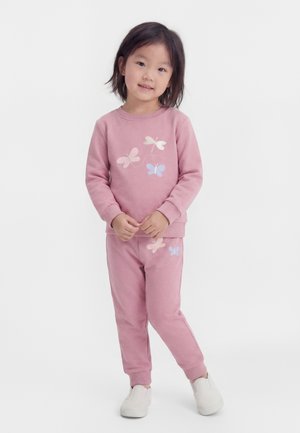 Jeune fille debout, portant un sweat-shirt rose et un pantalon avec des imprimés de papillons, et des chaussures blanches à enfiler, souriant légèrement.