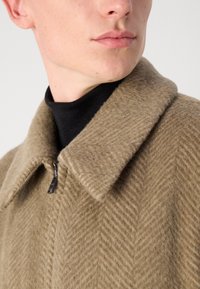 Beige Wollsakko im Fischgratmuster, mit großem Kragen und Reißverschluss, einer strukturierten Oberfläche und einem enganliegenden schwarzen Rollkragenpullover darunter.