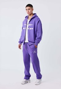 Joven de pie vistiendo un chándal púrpura PXLP con chaqueta con capucha y cremallera, pantalones a juego, camiseta blanca y zapatillas blancas sobre un fondo blanco.