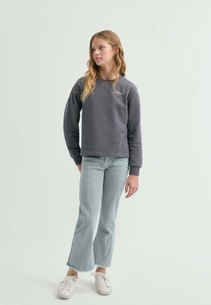 Mujer joven de pie con sudadera gris oscuro, jeans acampanados cortos azul claro y zapatillas blancas, mirando hacia un lado.