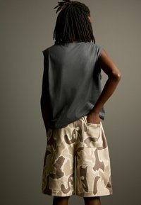 Mouwloos grijs T-shirt gecombineerd met beige camouflage shorts tot de knie, voorzien van een zijzak. De stof lijkt zacht en heeft een relaxte pasvorm.