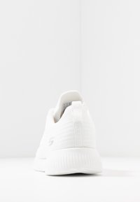 En ensam vit sneaker sedd från baksidan, som visar ribbad sula, texturerad ovandel och dragflik på en vit yta.