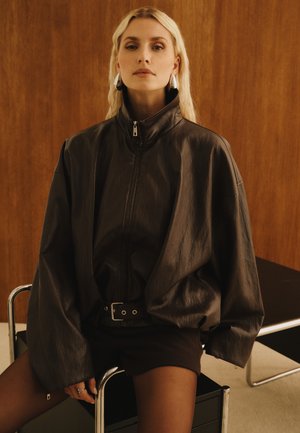 Mujer rubia sentada en una silla con una chaqueta de cuero negra y holgada, pantalones cortos negros, pendientes de aro plateados y anillos frente a una pared de madera.