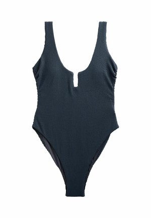 Femeie într-un costum de baie întreg, texturat și de culoare închisă, stând pe o plajă cu nisip, cu valurile oceanului pe fundal.