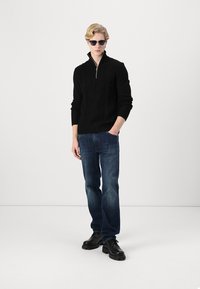 Pull en maille câble noir avec un zip quart, associé à un jean en denim bleu et des chaussures noires. Le mannequin porte des lunettes de soleil et se tient sur un fond uni.