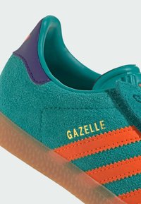 Green suede sneaker with orange stripes and a yellow "Gazelle" logo. Features a teal lining and a purple heel accent. Clear rubber sole.