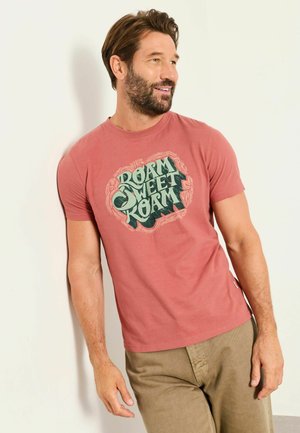 Mann trägt ein ausgewaschenes rotes T-Shirt mit dem Text "Roam Sweet Roam", lehnt an einer weißen Wand, schaut zur Seite und lächelt.