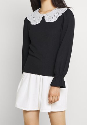 Woman wearing a black long-sleeve top with a white lace collar and white high-waisted shorts, standing against a plain background.