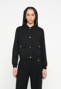 Jachetă scurtă neagră cu guler rotund, nasturi capse argintii, două buzunare frontale și manșete striate. Țesătură simplă, texturată, cu un design ajustat.