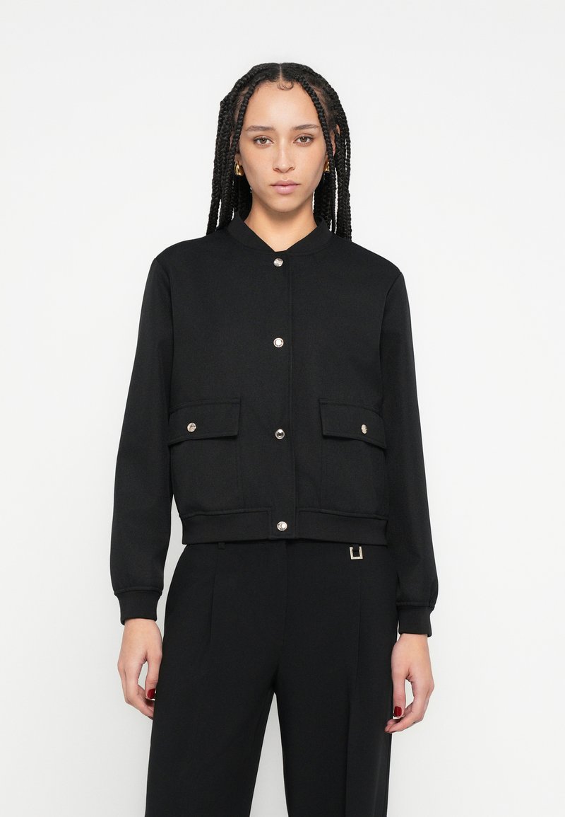 Jachetă scurtă neagră cu guler rotund, nasturi capse argintii, două buzunare frontale și manșete striate. Țesătură simplă, texturată, cu un design ajustat.