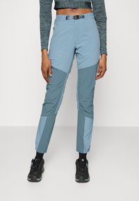 Ljusblå utomhusbyxor med mörkare blå accenter, en figurnära passform, elastiska mudd och sidofickor med dragkedja; tillverkade av ett slitstarkt material.