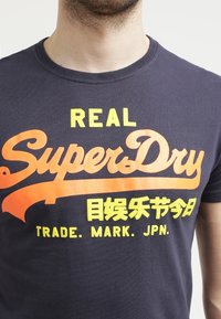 Námořnická modrá bavlněná trička s výrazným oranžovým a žlutým textem. Text obsahuje "REAL SuperDry" a další značkové detaily v různých písmách.