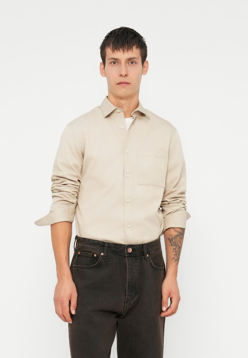 Μπεζ πουκάμισο με κουμπιά, φτιαγμένο από βαμβάκι, με μακριά μανίκια και μία τσέπη στο στήθος, φορεμένο πάνω από μαύρα τζιν με ένα τατουάζ ορατό στον πήχη.