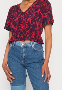 Rot-blaues Bluse mit floralem Muster, kurzen Ärmeln, V-Ausschnitt und Peplum-Saum, kombiniert mit hellblauen, hoch taillierten Jeans.