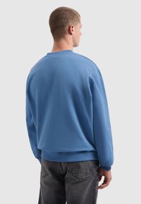 Man met kort haar, gekleed in een blauwe sweatshirt en donkergrijze jeans, staande met de rug naar de camera tegen een effen lichtgrijze achtergrond.