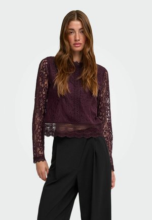 Femme portant un top en dentelle violet foncé à manches longues et un pantalon noir taille haute à jambes larges, debout devant un fond uni.