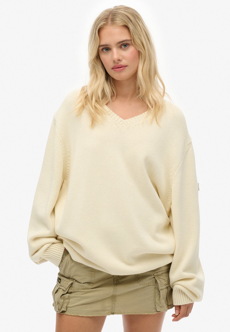 Blondynka w oversize'owym kremowym swetrze z dzianiny i krótkiej oliwkowej spódnicy cargo, stojąca na tle jednolitej białej ściany.