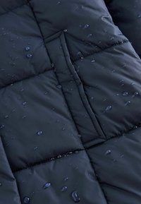 Vodootporni tamnoplavi puffer kaput od tkanine s kvadratnim šavovima i vidljivim kapljicama vode na površini, naglašavajući njegovu zaštitnu strukturu.